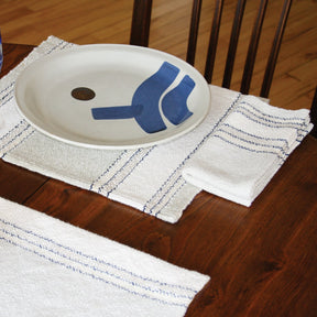 A wooden dining table is set with a white stoneware dish featuring an abstract, blue geometric design. The plate rests on the Rustic Mats from Halcyon Yarn, a white placemat with blue stitching, and matching white napkins with blue stitching are positioned neatly to the right and below the placemat.