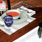 A table set with a bowl of tomato soup on a white plate, stoneware dishes surrounding it, and utensils resting on Rustic Mats placemats by Halcyon Yarn. A crystal glass with blue liquid adds a burst of color, while a teapot peeks in from the right. Overhead, the Adobe PDF logo lingers in the top left corner.