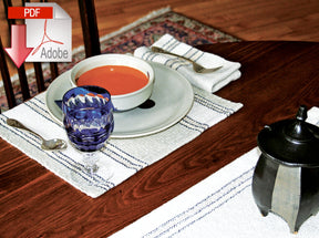 A table set with a bowl of tomato soup on a white plate, stoneware dishes surrounding it, and utensils resting on Rustic Mats placemats by Halcyon Yarn. A crystal glass with blue liquid adds a burst of color, while a teapot peeks in from the right. Overhead, the Adobe PDF logo lingers in the top left corner.