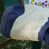 A knitted blanket with a light beige center and dark blue edges is draped over the arm of a green outdoor chair. Nearby, the stylish Casco Bay Tote by Halcyon Yarn adds a touch of elegance to the scene, while in the background, there is a partially visible book with illustrated colorful shapes on the cover.