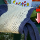 A knitted sweater, half in cream and half in purple, is draped over a green table. In the background, a box of vine-ripened tomatoes and corn can be seen alongside a stylish Casco Bay Tote from Halcyon Yarn. A red Adobe PDF icon with a downward arrow indicating "download" is placed on top of the image.