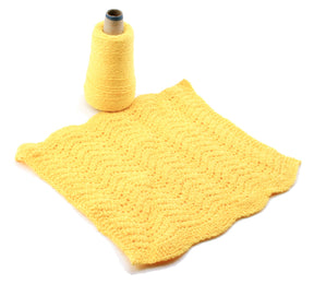 A vibrant yellow crocheted square with a delicate wavy pattern lies flat on a pristine white surface, paired with a matching yellow yarn spool from Halcyon Yarn's Washcloths / Dishcloths Sampler, standing upright beside it. This exquisite piece would make an ideal gift or serve as part of detailed washcloth patterns.