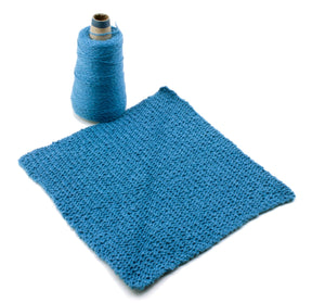 A blue knitted square with a textured pattern is laid flat on a white surface next to a tall spool of Halcyon Yarn. The spool is nearly as tall as the square is wide—perfect for creating intricate patterns that make an elegant gift using the Washcloths / Dishcloths Sampler by Halcyon Yarn.