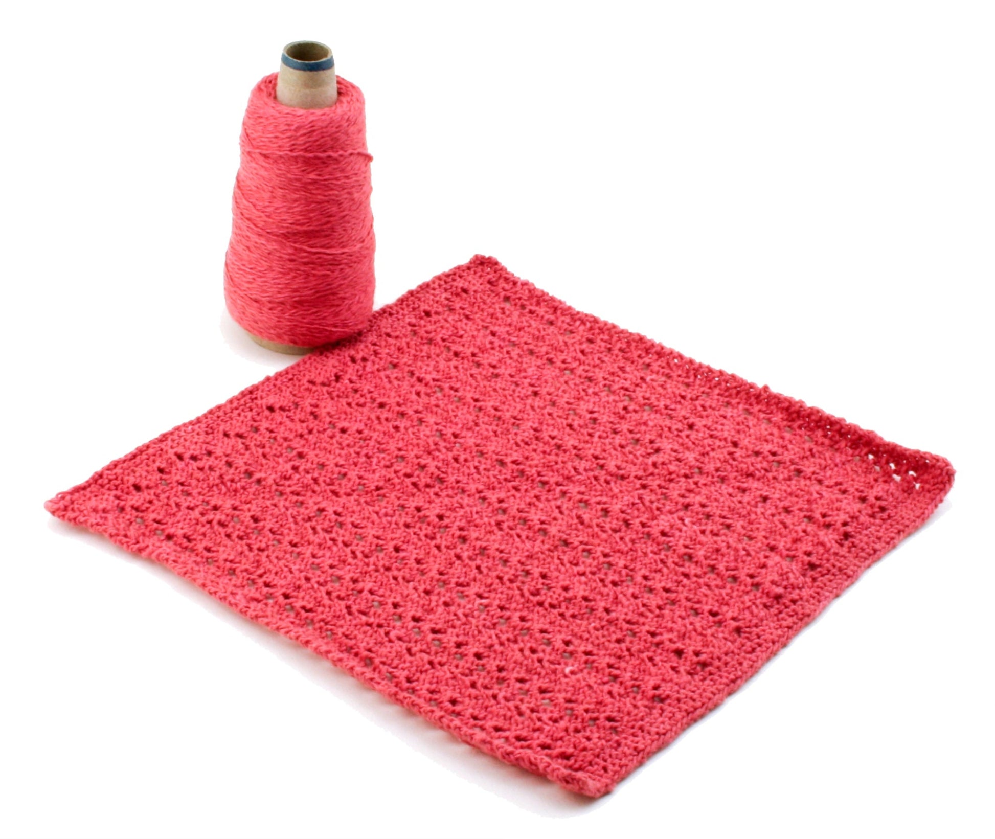 A pink crocheted square, ideal for washcloth patterns, is laid flat next to a spool of Halcyon Yarn Casco Bay Sport in matching pink. The thick, textured yarn beautifully enhances the lacy, intricate pattern of the square. Both items are perfectly displayed on a white background, showcasing the Washcloths/Dishcloths Sampler by Halcyon Yarn.