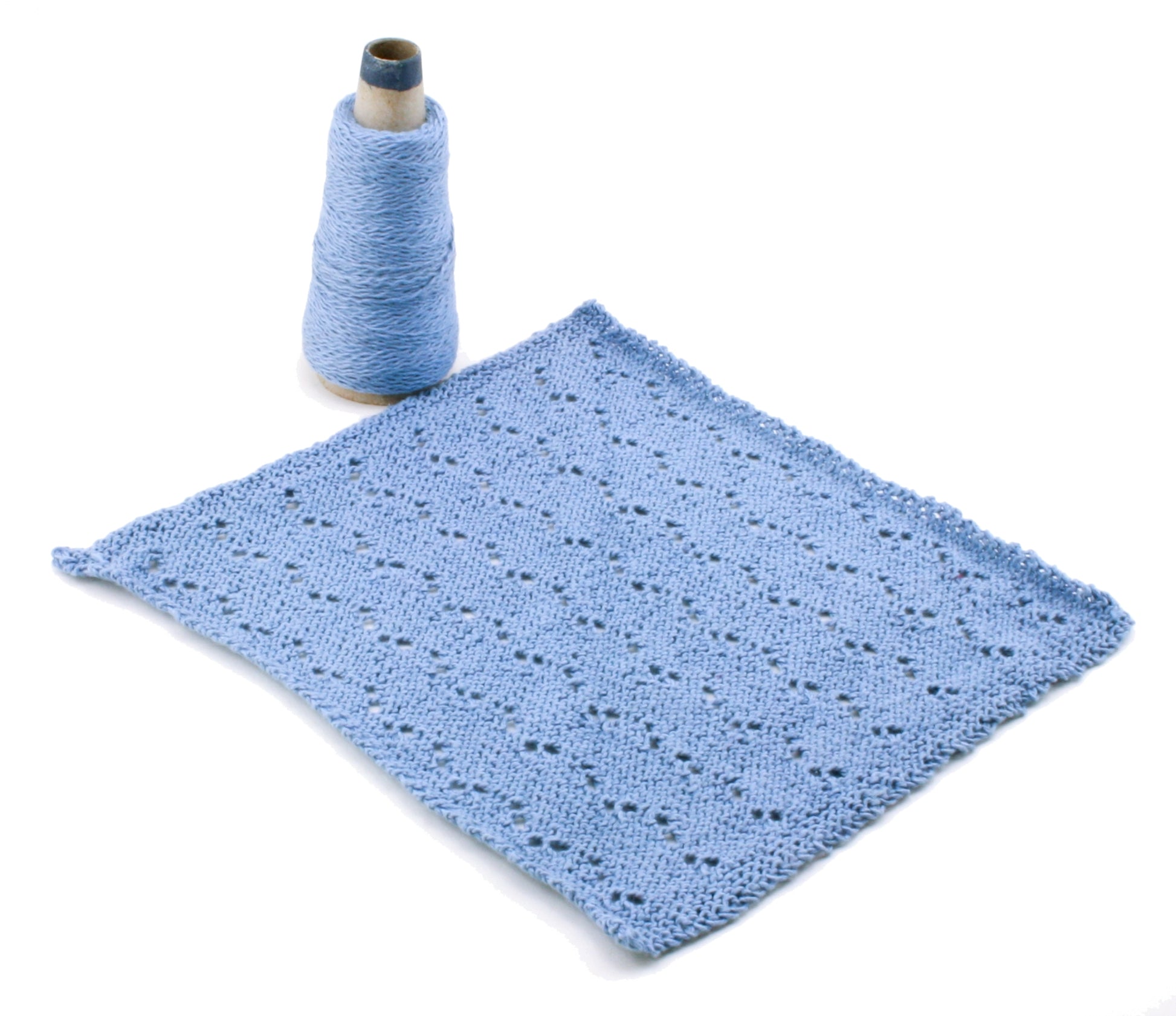 A textured, light blue knit square is laid out flat beside a cone of matching Casco Bay Sport cotton yarn. The knitting pattern features small eyelet holes arranged in diagonal rows, creating a decorative design ideal for Halcyon Yarn's Washcloths / Dishcloths Sampler download.