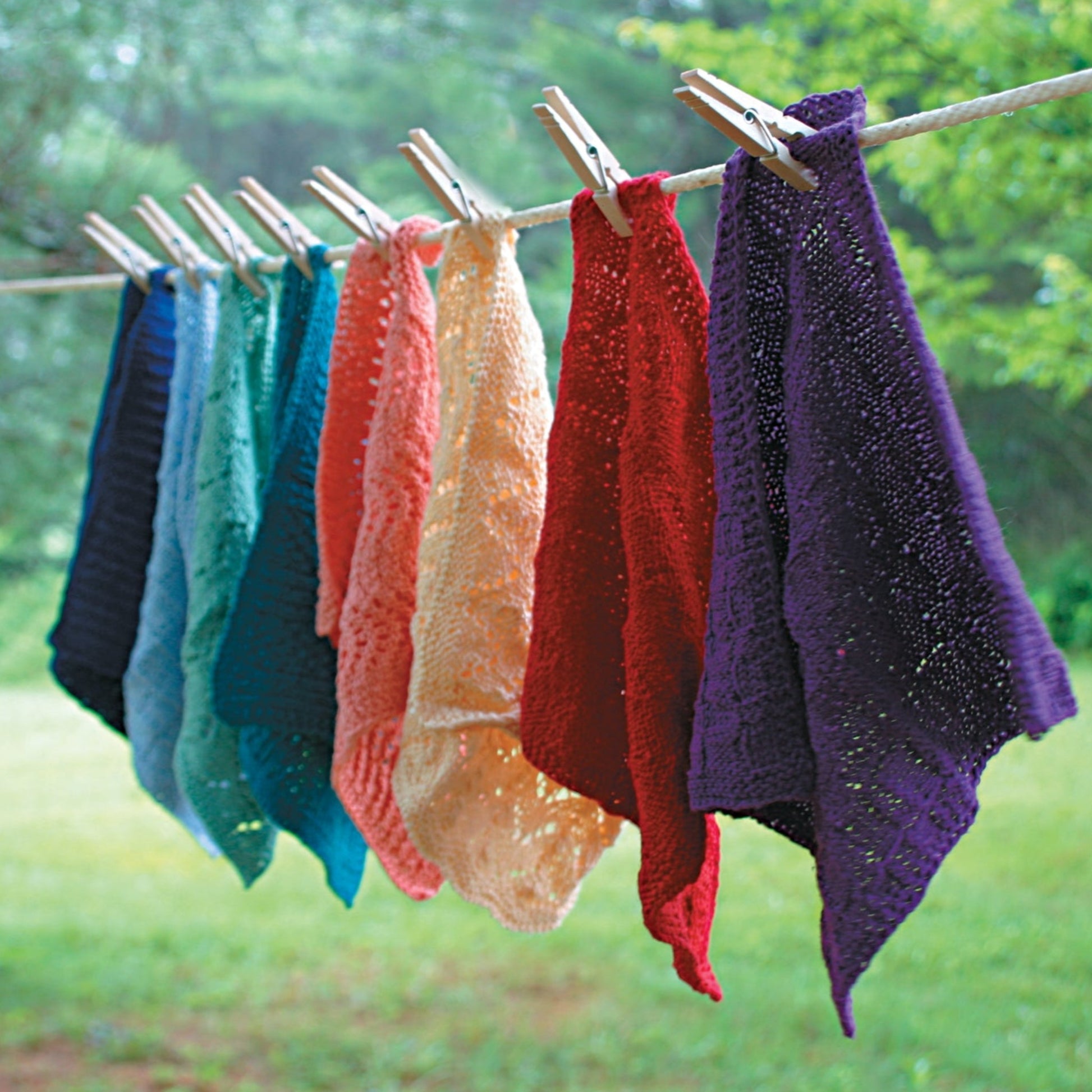 A row of colorful washcloths and dishcloths from the Halcyon Yarn Washcloths / Dishcloths Sampler are hanging on a clothesline with wooden clothespins. The vibrant cloths in blue, teal, green, orange, yellow, red, and purple make perfect gifts. The background features a lush green outdoor setting.