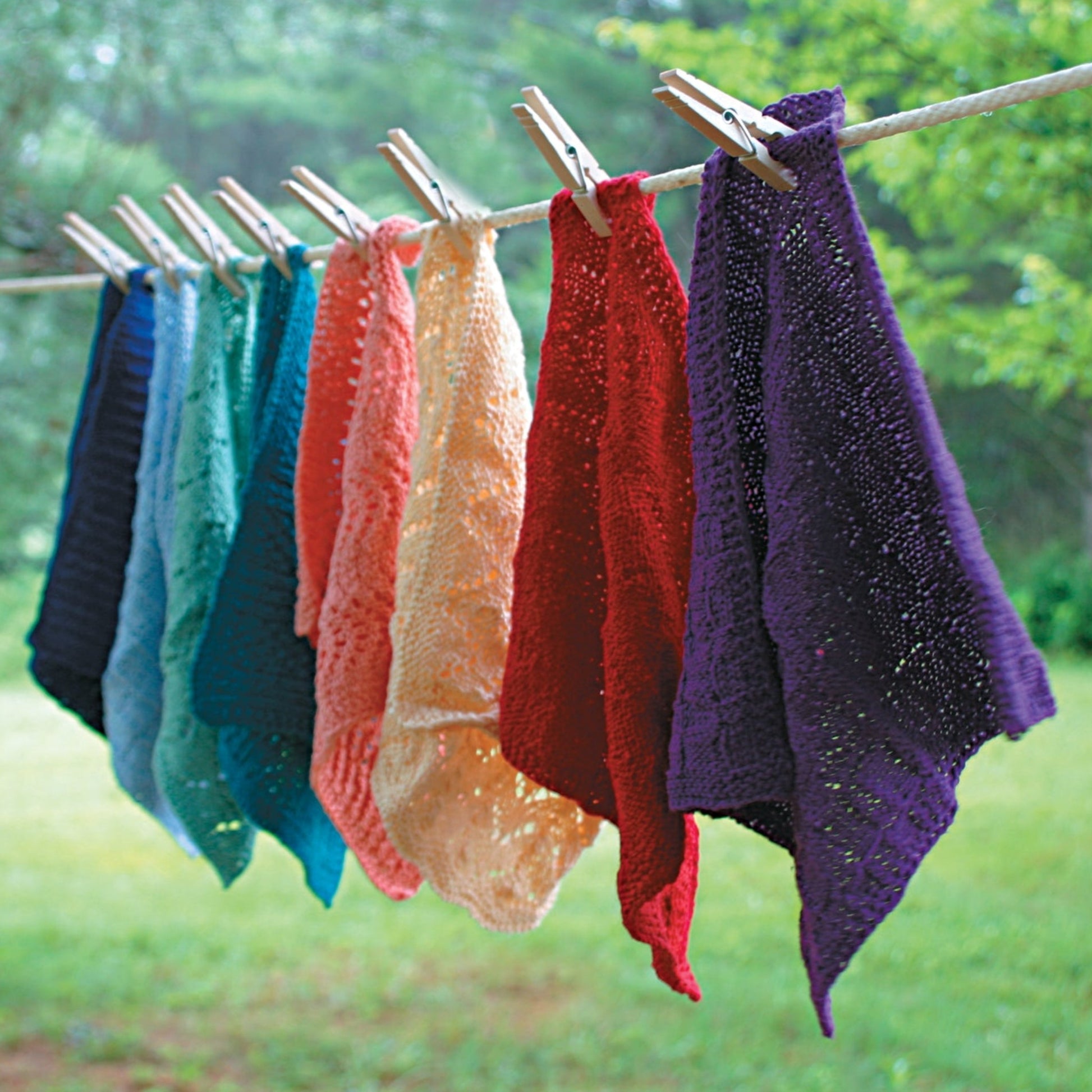 A row of the vibrant Halcyon Yarn Washcloths / Dishcloths Sampler in shades of blue, green, turquoise, orange, yellow, red, and purple hang on a clothesline with wooden clothespins. Made from soft cotton yarn and featuring intricate patterns, they sway gently against a backdrop of green trees and grassy area.