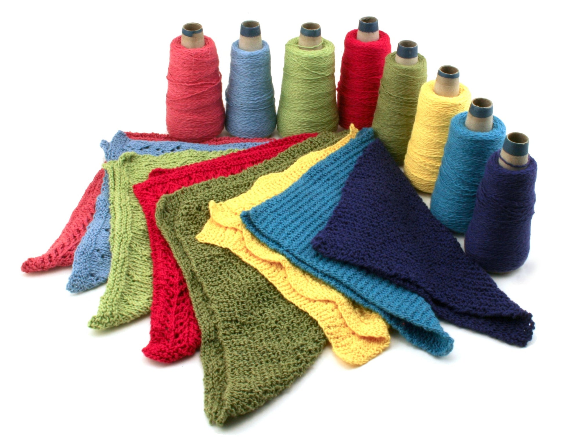 A variety of colorful yarn cones are lined up in the background, showcasing shades of red, green, yellow, blue, and purple. In front, there are knitted fabric swatches in matching colors from the Halcyon Yarn Washcloths / Dishcloths Sampler set, elegantly displayed in a fan-like arrangement on a white surface—perfect for crafting unique wash cloth patterns as a thoughtful gift.