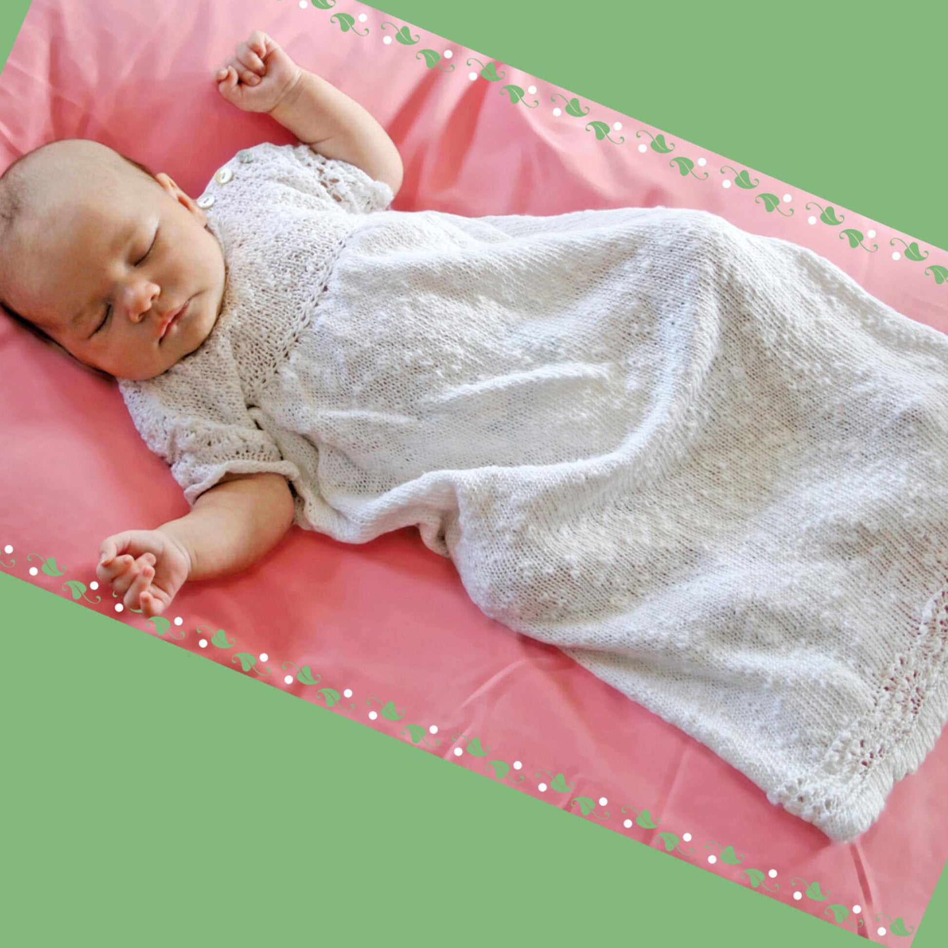 A sleeping baby is lying on a pink surface, swaddled in a Halcyon Yarn Christening Gown. The background is green with a decorative floral border. The baby's arms are positioned gently beside them, with hands slightly open, embracing the beauty of new life and the gown's beautiful texture.