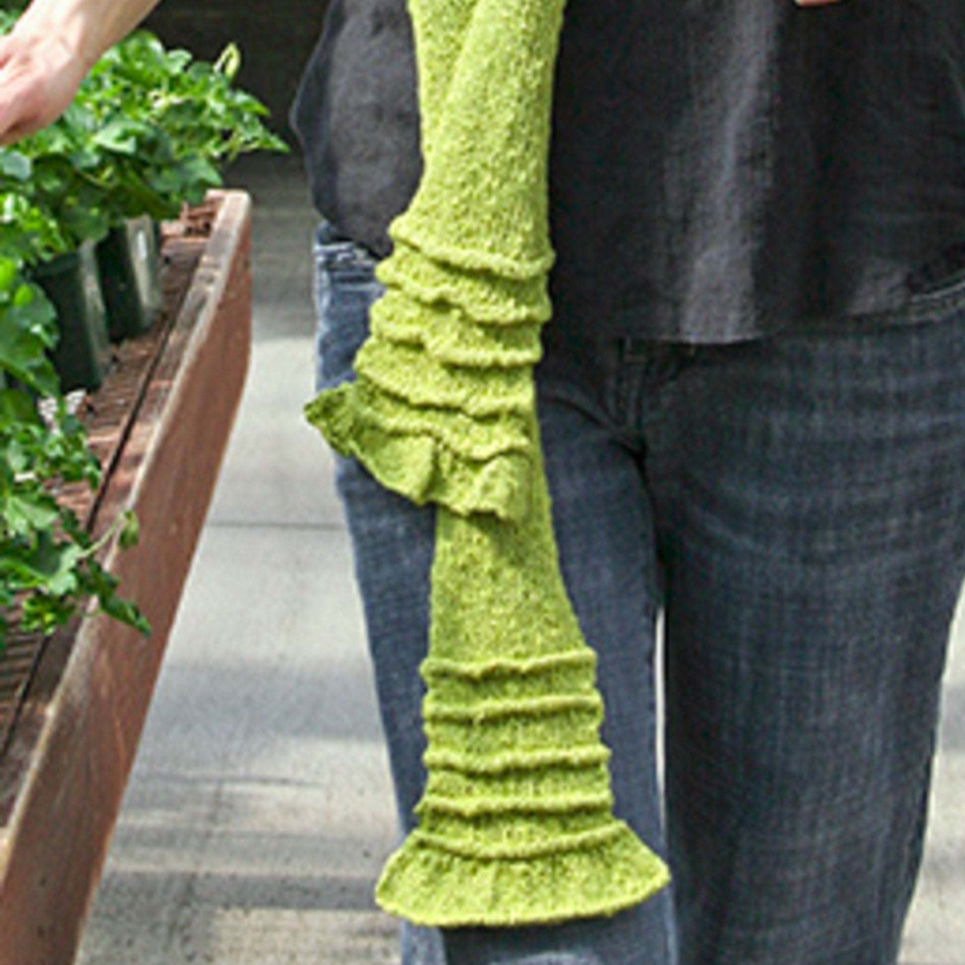 A person wearing a black shirt and blue jeans is holding Halcyon Yarn's Accordion Accent Scarf in green, which features a slight ruffle. They are standing next to a wooden table adorned with potted plants. The image captures the lower half of their body and the edge of the table, showcasing how the Accordion Accent Scarf can enhance any outfit.