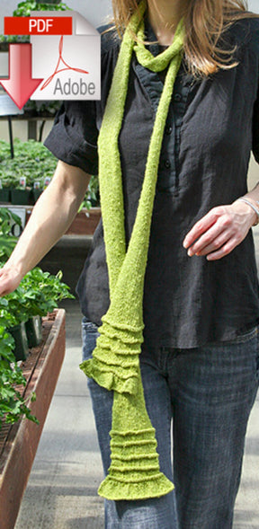 A person dressed in a black blouse and blue jeans is sporting an Accordion Accent Scarf from Halcyon Yarn, featuring a slight ruffle that enhances any outfit. They're standing in what seems to be a garden center, surrounded by potted plants. An Adobe PDF icon can be seen superimposed in the upper left corner of the image.