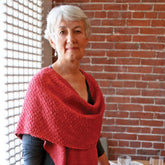 An elderly person with short, gray hair is wearing a red Woven Cowl Wrap from Halcyon Yarn over a black top. They stand against a brick wall beside a glass-bricked window, with a table and glass visible in the foreground. The cozy scene perfectly captures the essence of Halcyon's Signature Block Island Blend.