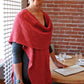 An older woman with short gray hair stands indoors near a table set for a meal. She is wearing a Woven Cowl Wrap from Halcyon Yarn over her black dress, adding warmth and elegance to her ensemble. The background features a brick wall and a mesh-like room divider. She is holding a menu and looking at the camera.