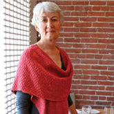 An older woman with short gray hair stands indoors against a brick wall. She is wearing a Woven Cowl Wrap from Halcyon Yarn over a black top and holds an expression of calm confidence. The room has a window with a grid pattern allowing light to gently illuminate the space.