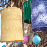 Lumen-patterned lampshades by Twist Collective in yellow, green, and purple hang from a tree, beautifully crafted with delicate fingering weight yarn. In the blurred background, additional lantern covers and lush foliage enhance the enchanting scene.