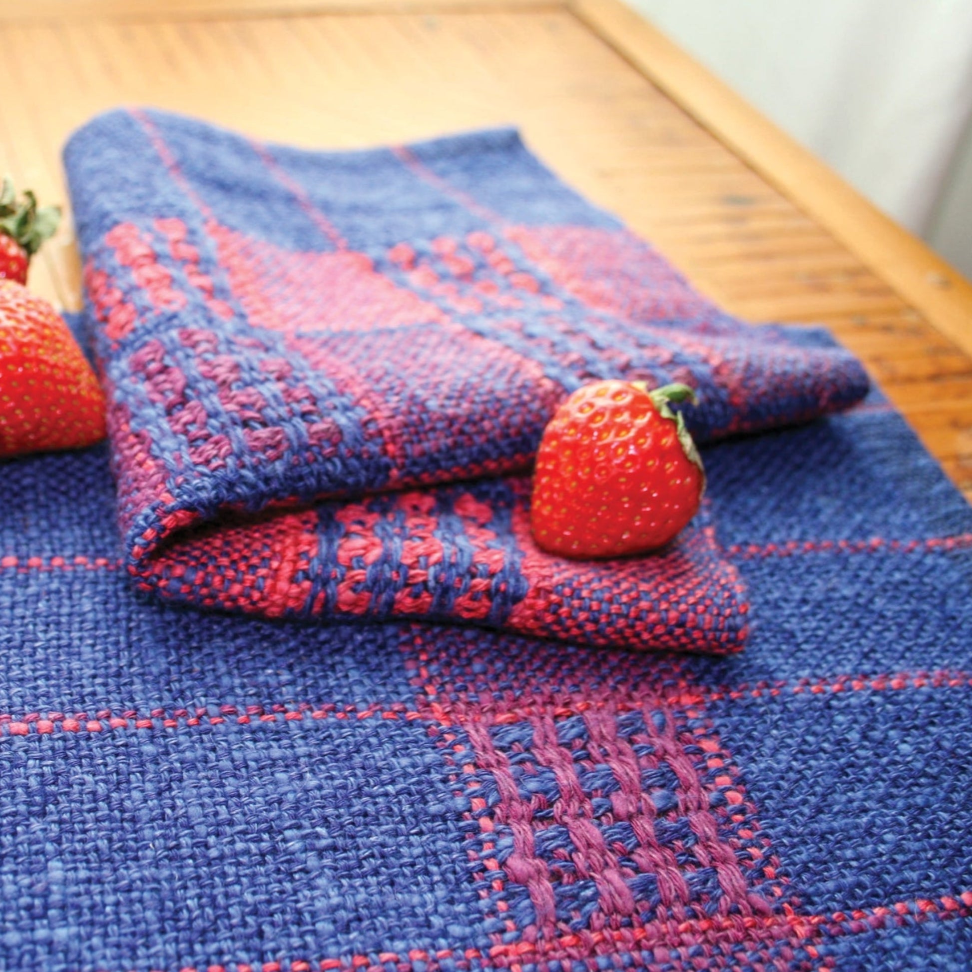 A folded blue and red handwoven textile with a checked pattern lies on a wooden surface. Two fresh strawberries rest on top of the textile, adding a pop of natural color to the scene, evocative of the Seguin Sunset Table Set by Halcyon Yarn.