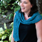 A woman stands outdoors, smiling, with a Heirloom Lace Signature Scarf from Halcyon Yarn in light blue draped around her neck. She has medium-length dark hair and is wearing a sleeveless black top. Green foliage and a tree trunk are visible in the background.