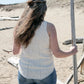 The person with long, wavy brown hair, wearing the Sweet Jane Tank by Halcyon Yarn and blue jeans, stands on a sandy beach. They hold a tall stick in their left hand and face away from the camera, with scattered logs and dunes visible in the background.