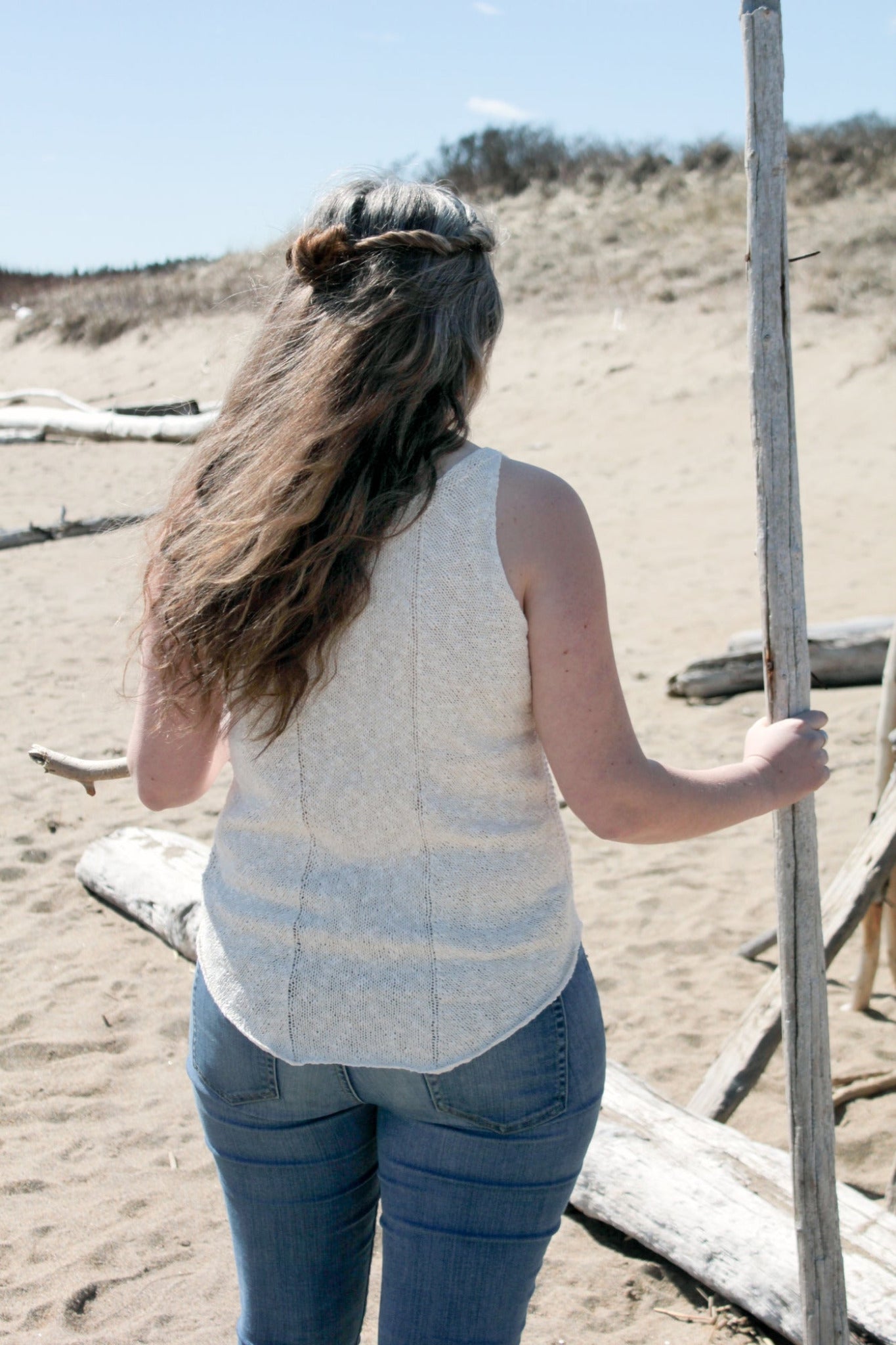 The person with long, wavy brown hair, wearing the Sweet Jane Tank by Halcyon Yarn and blue jeans, stands on a sandy beach. They hold a tall stick in their left hand and face away from the camera, with scattered logs and dunes visible in the background.