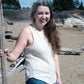 A woman with long brown hair and a white streak is smiling at the camera, standing on a sandy beach. She is wearing the Halcyon Yarn Sweet Jane Tank and blue jeans, holding a long wooden stick. Several logs and trees are in the background under a blue sky with scattered clouds.