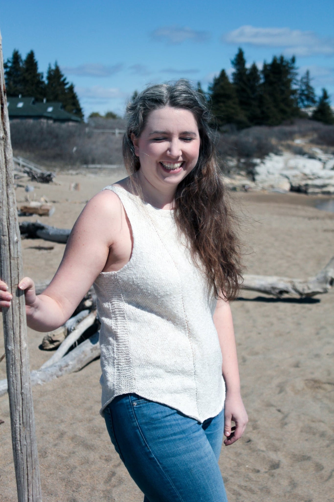 A woman with long brown hair and a white streak is smiling at the camera, standing on a sandy beach. She is wearing the Halcyon Yarn Sweet Jane Tank and blue jeans, holding a long wooden stick. Several logs and trees are in the background under a blue sky with scattered clouds.