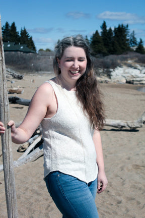 A woman with long, wavy hair stands on a sandy beach, smiling at the camera. She is wearing the Sweet Jane Tank - Pattern Download by Halcyon Yarn and blue jeans, holding a long stick with one hand. The beach has scattered driftwood, and trees are visible in the background under a partly cloudy sky.