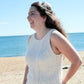 A woman with long, wavy hair is smiling and looking off to the side while standing on a sandy beach. She is wearing the Sweet Jane Tank by Halcyon Yarn, which features a rustic texture. The ocean and clear blue sky are visible in the background.