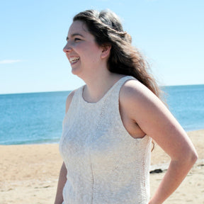 A person with long hair is standing on a beach, smiling and looking towards their left. They are wearing the Halcyon Yarn Sweet Jane Tank - Pattern Download, featuring stylish bias panels. The background includes a sandy shore and a calm, blue ocean under a clear sky.