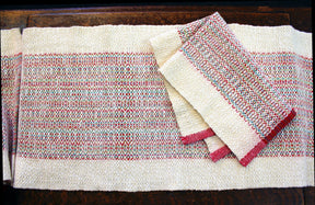 A Homeward Bound Table Runner and Placemat Set from Halcyon Yarn, crafted from natural fibers and featuring intricate patterns in red, green, and beige, lies on a dark wooden surface. The placemats are neatly folded on top of the table runner, showcasing their matching design.