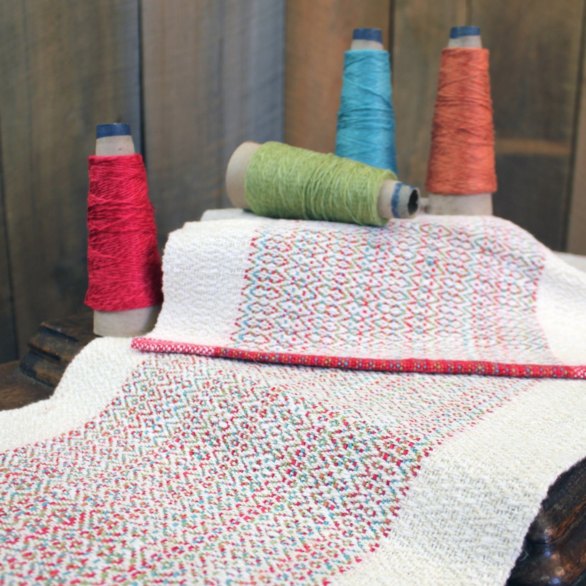 A sophisticated white fabric with intricate multicolored patterns from the Homeward Bound Table Runner & Placemat Set by Halcyon Yarn is draped over a wooden surface. Four spools of thread in red, green, blue, and orange colors are perched behind the fabric, adding a splash of color to the scene. The use of natural fibers enhances the elegance of this beautiful display.