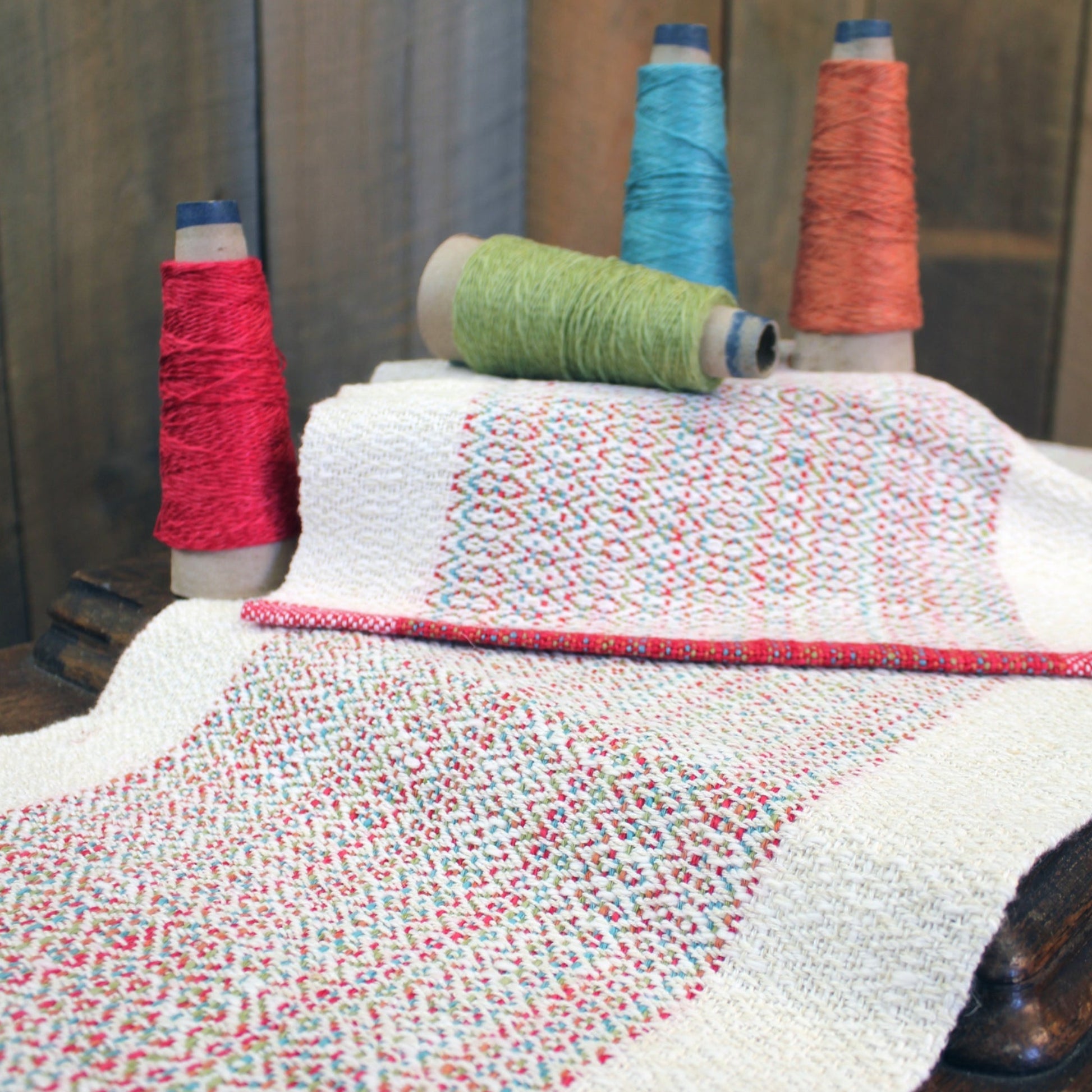 Displayed on a wooden surface is the Homeward Bound Table Runner & Placemat Set by Halcyon Yarn, featuring a sophisticated fabric with a colorful and intricate design. Alongside the fabric are four spools of thread in red, green, turquoise, and orange shades. The threads harmonize perfectly with the colors in the textile's detailed pattern, highlighting its high absorbency thanks to its natural fibers.