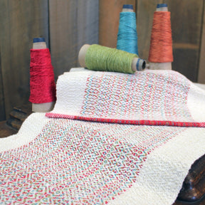 Displayed on a wooden surface is the Homeward Bound Table Runner & Placemat Set by Halcyon Yarn, featuring a sophisticated fabric with a colorful and intricate design. Alongside the fabric are four spools of thread in red, green, turquoise, and orange shades. The threads harmonize perfectly with the colors in the textile's detailed pattern, highlighting its high absorbency thanks to its natural fibers.