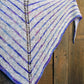 Introducing the Electric Avenue Shawlette by Halcyon Yarn, a hand-knitted summer accessory adorned with alternating stripes of white and various shades of blue and purple. This exquisite shawl features a central lace pattern running from top to bottom, crafted from Signature Hand Dyed Silk Noil. For an elegant presentation, the shawlette is displayed against a wooden background.