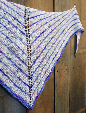 Introducing the Electric Avenue Shawlette by Halcyon Yarn, a hand-knitted summer accessory adorned with alternating stripes of white and various shades of blue and purple. This exquisite shawl features a central lace pattern running from top to bottom, crafted from Signature Hand Dyed Silk Noil. For an elegant presentation, the shawlette is displayed against a wooden background.