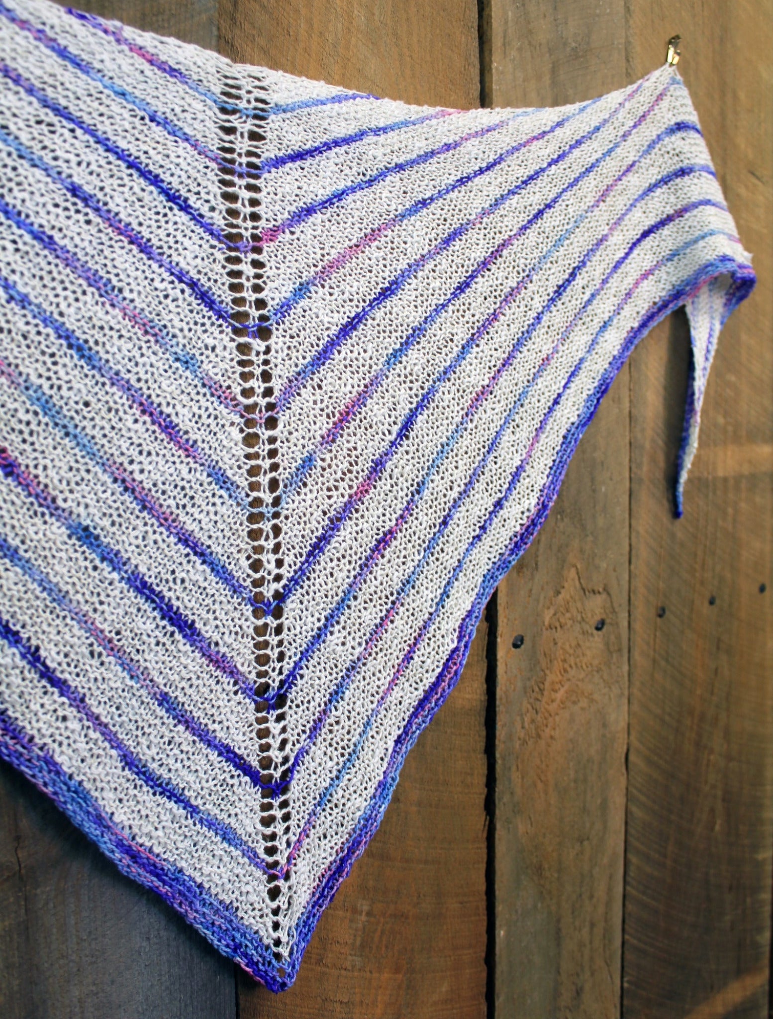 The Electric Avenue Shawlette by Halcyon Yarn, showcasing a striped pattern in various shades of blues, purples, and whites, hangs against a wooden background. Crafted from Signature Block Island Blend, the design features a central eyelet row running down the middle that separates the pattern symmetrically on both sides.