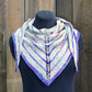 An Electric Avenue Shawlette by Halcyon Yarn in stunning shades of white, purple, and blue is draped on a black mannequin. The triangular summer shawlette features an intricate lace pattern down the center and is elegantly displayed against a wooden background.