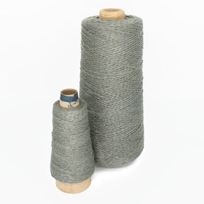 Two spools of gray thread stand on a white background. The larger spool, on the right, is Halcyon Yarn's Block Island Blend | Large Cone and has thicker thread. The smaller spool, on the left, contains thinner thread. Both spools have wooden bases.