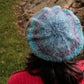 A person in a red sweater is viewed from behind, donning a Relaxed Beret by Halcyon Yarn made of self-striping yarn in various shades of blue and grey. They are standing outdoors on grass with rocks in the background.