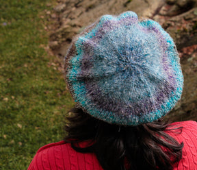 A person in a red sweater is viewed from behind, donning a Relaxed Beret by Halcyon Yarn made of self-striping yarn in various shades of blue and grey. They are standing outdoors on grass with rocks in the background.