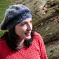 A woman wearing a Relaxed Beret from Halcyon Yarn, knitted with vibrant self-striping yarn, and a red sweater stands outdoors, looking slightly upward with a smile. She is in front of a rock formation with a natural, earthy background.