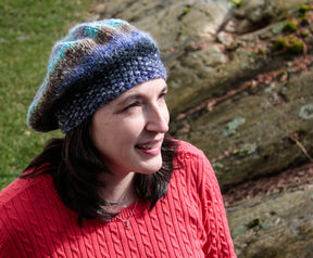 A woman wearing a Relaxed Beret from Halcyon Yarn, knitted with vibrant self-striping yarn, and a red sweater stands outdoors, looking slightly upward with a smile. She is in front of a rock formation with a natural, earthy background.