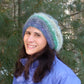 A person with long dark hair is wearing the Relaxed Beret from Halcyon Yarn, made from self-striping yarn in green, gray, and blue. Paired with a blue jacket, they are standing outdoors in front of a backdrop of evergreen trees, smiling at the camera.