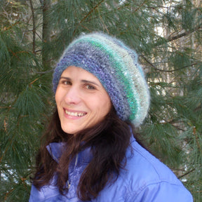 A woman with long dark hair smiles while wearing the Relaxed Beret, a pattern available for download from Halcyon Yarn, and a blue jacket. She stands outside with green pine trees in the background.