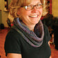 A woman with short blonde hair and glasses smiles warmly at the camera. She is wearing a black shirt and the "Cowl Two Ways" from Halcyon Yarn, a multicolored knit scarf made from variegated yarn. The background appears to be a cozy indoor setting with wooden floors and shelves.