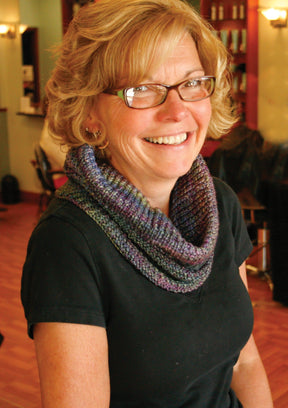 A woman with short blonde hair and glasses smiles warmly at the camera. She is wearing a black shirt and the "Cowl Two Ways" from Halcyon Yarn, a multicolored knit scarf made from variegated yarn. The background appears to be a cozy indoor setting with wooden floors and shelves.