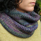 Close-up of a person wearing the "Cowl Two Ways" by Halcyon Yarn, crafted from variegated yarn featuring shades of blue, purple, and green. The textured surface adds a snug feel, perfectly complementing their light green sweater. The background is blurred with greenery.