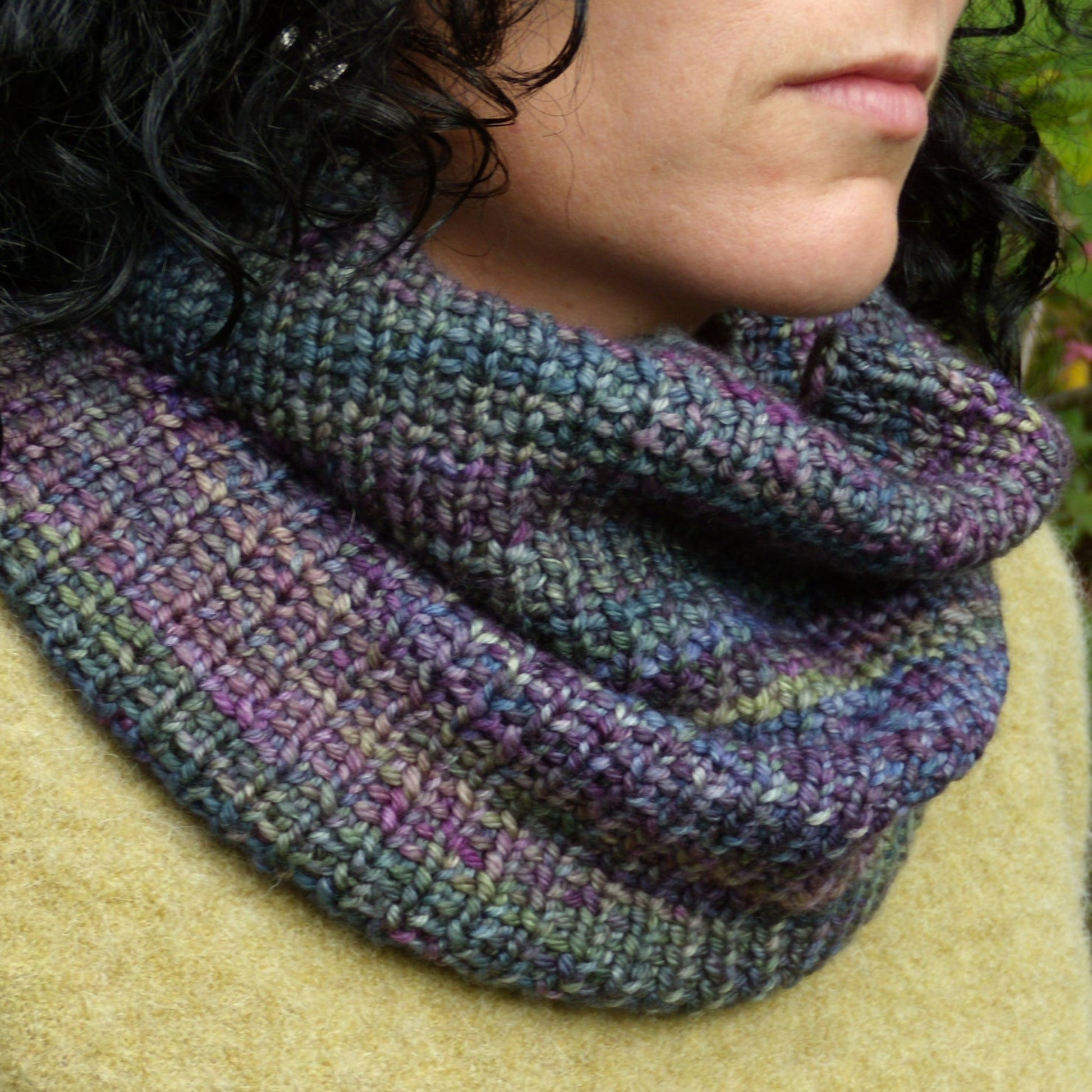 A close-up of a person wearing the "Cowl Two Ways" from Halcyon Yarn, a textured, multicolored knit cowl scarf made with variegated yarn. The quick-knit cowl features hues of purple, green, and blue. The person has short, curly dark hair and is dressed in a light brown sweater. Only the lower part of their face and neck are visible.