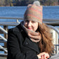 A woman sits at an outdoor table near a body of water on a sunny day. She is wearing a black coat and the "Telephone Line Set, Hat & Cowl" from Halcyon Yarn, featuring a multicolored knit hat with a pom-pom made from DK Wool and a matching scarf around her neck. The background features trees and blue water under a clear sky.