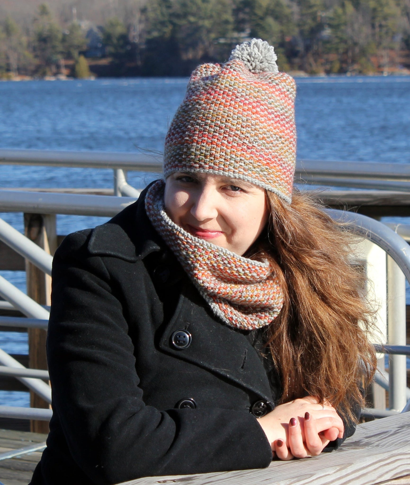 A woman sits at an outdoor table near a body of water on a sunny day. She is wearing a black coat and the "Telephone Line Set, Hat & Cowl" from Halcyon Yarn, featuring a multicolored knit hat with a pom-pom made from DK Wool and a matching scarf around her neck. The background features trees and blue water under a clear sky.