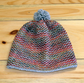 A hat from the Telephone Line Set, Hat & Cowl collection by Halcyon Yarn, crafted from DK Wool and featuring a multicolored pattern of red, green, and gray yarn with a light gray pom-pom on top, is displayed on a wooden surface.